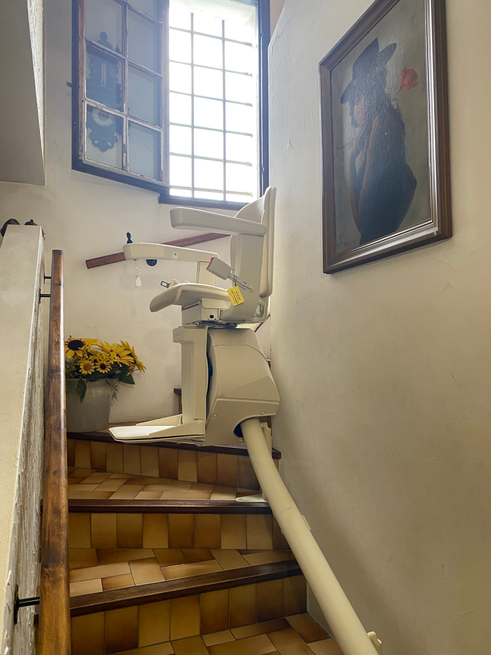 Monte-escalier replié en haut d'un escalier carrelé, près d'une fenêtre et d'un bouquet de tournesols